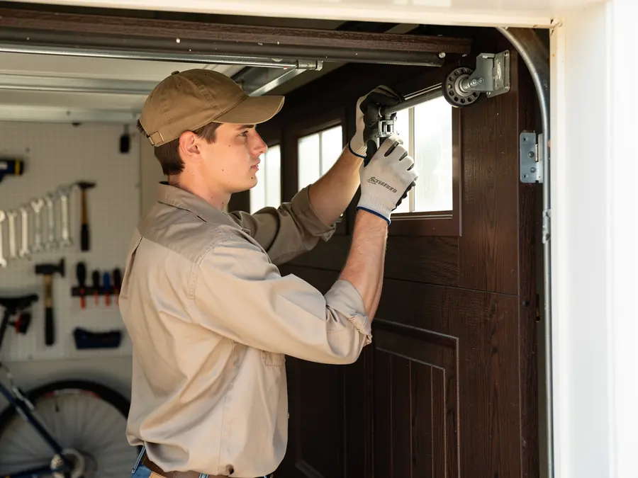 A technician in a cap and gloves works on a brown garage door.